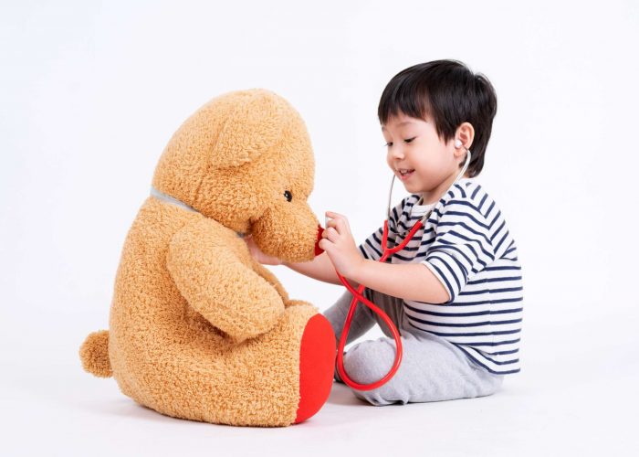 Asian cute boy playing a doctor use stethoscope checking large teddy bear sitting on floor isolated on white background